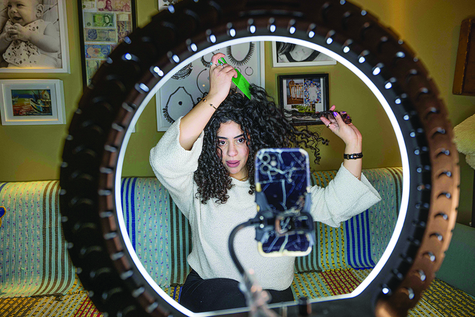 Mariam Ashraf combs her hair as she speaks before a phone on a tripod and lights during a live-stream.