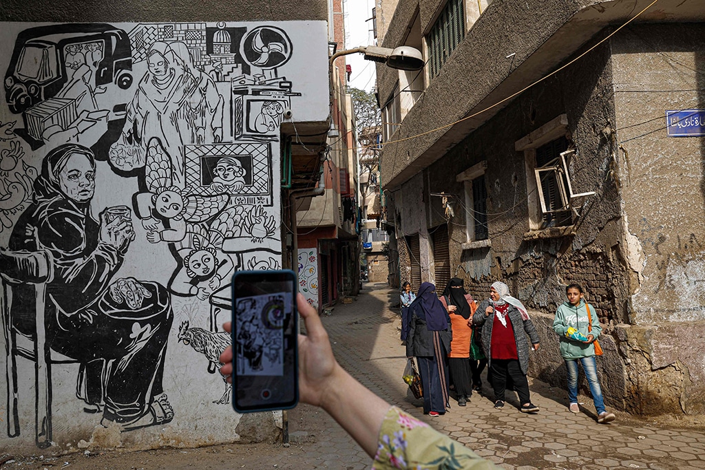People walk down the street as a woman uses a mobile phone to scan a mural to tell local residents about their heritage in a Cairo neighborhood.