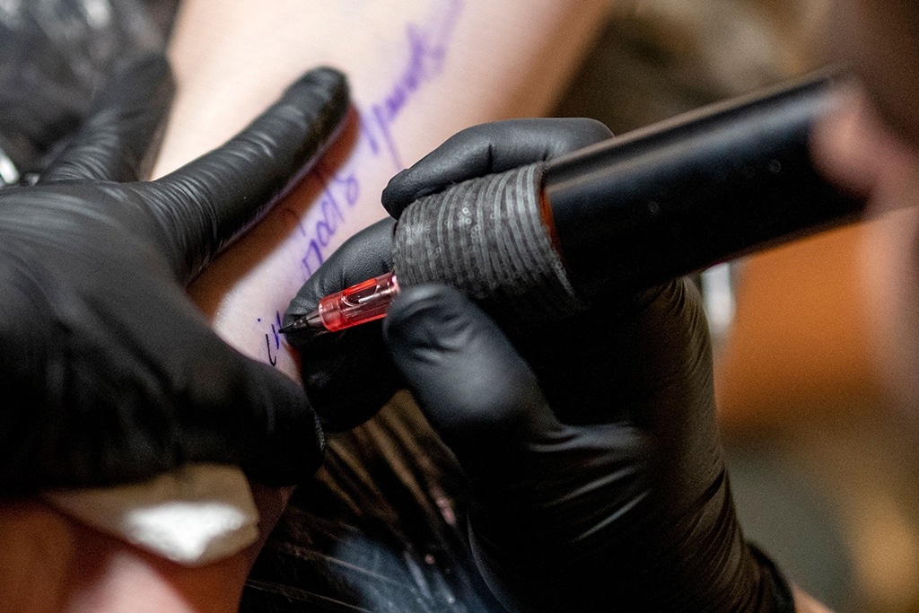 Tattoer Silas Becks curves a tattoo reading 'In God's hands..' to the arm of a woman.