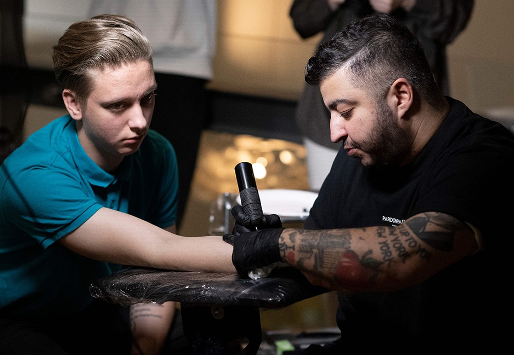 Tattoer Silas Becks curves a tattoo reading 'In God's hands..' to the arm of a woman.